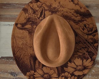 May include: A tan fedora-style hat with a brown fox illustration on the brim. The hat features a brown crown and a wide brim decorated with a fox, flowers, and foliage. The hat is likely made of felt or a similar material.