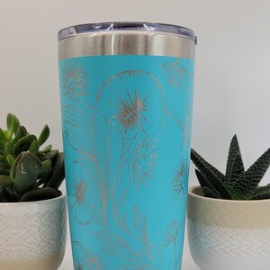 Könnte beinhalten: Ein türkisfarbener Edelstahlbecher mit einem silbernen Blumendesign. Der Becher hat einen Deckel und ist perfekt, um Getränke kalt oder warm zu halten.