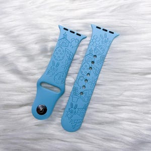 May include: Two light blue Apple Watch bands with Easter-themed designs. Each band features engraved images of Easter eggs, bunnies, chicks, flowers, and a church. The bands are made of a flexible material and are designed to fit an Apple Watch.