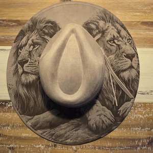 May include: A light grey fedora hat with a wide brim, featuring a detailed illustration of two lion faces in dark grey. The hat has a decorative string detail on the side. The hat is resting on a wooden surface.