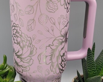 May include: A pink insulated tumbler with a floral design in silver. The tumbler has a handle and a white lid. The design features large peony flowers and leaves. The tumbler is next to two small potted plants.