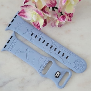 May include: Two light blue watch bands with airplane designs. The bands feature various airplane illustrations, propellers, and engine details. The bands are made of a flexible material and are designed to fit a smartwatch. The background includes pink and white flowers.