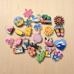 Könnte beinhalten: Eine Sammlung farbenfroher Charms zum Thema Tropen. Designs umfassen eine Wassermelonen-Scheibe, Eis am Stiel, Palmen, einen Surfer, einen Flamingo in einem Pool-Float, eine Margarita und eine Muschel. Die Charms sind in verschiedenen Formen und Größen.