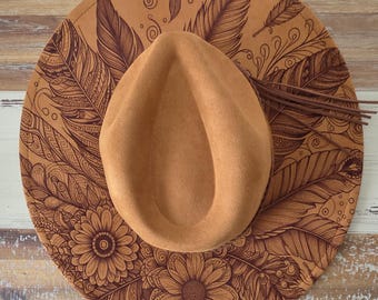 May include: A tan-colored hat with a wide brim, featuring a detailed feather and floral design etched into the brim. The hat has a teardrop-shaped crown and a leather fringe detail. The hat is displayed on a wooden surface.