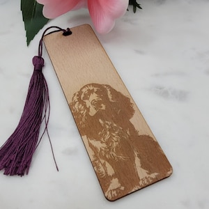 Puede incluir: Marcapáginas de madera con una imagen grabada de un perro, un Cocker Spaniel. Tiene una borla morada oscura. El marcapáginas está hecho de madera clara. La imagen del perro es detallada.