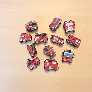Op de afbeelding: Een verzameling van elf kleine, rode brandweerwagen-schoenbedels. Elke bedel is voorzien van een gedetailleerde afbeelding van een brandweerwagen met zwarte wielen, witte accenten en diverse ladderconfiguraties voor in schoenen.