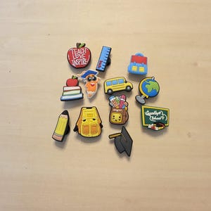 May include: A collection of school-themed shoe charms featuring a red apple with the text TEACH LOVE INSPIRE, a blue ruler, a backpack, a stack of books, a pencil character, a yellow school bus, a globe, a yellow bag, a chalkboard with Goodbye School.