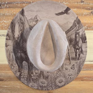 May include: A light gray felt hat featuring a detailed dark brown laser-engraved design on the brim. The artwork depicts two horses, a soaring eagle, mountain peaks, a forest, and various wildflowers and butterflies across the entire surface.