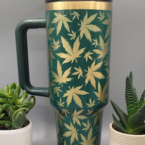 Puede incluir: Una taza de viaje verde con un patrón de hojas de marihuana doradas. La taza tiene una tapa negra y un asa.