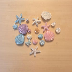 Puede incluir: Una colección de cabujones decorativos dispuestos en forma de corazón, con conchas, estrellas de mar, perlas, una mariposa y piezas en forma de corazón en tonos pastel, azul, rosa, blanco y acabados iridiscentes.