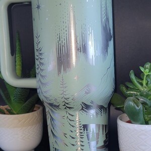May include: A mint green travel mug with a silver mountain and forest design. The mug has a handle, a white lid, and is surrounded by small potted plants. The mug is approximately 9 inches tall.
