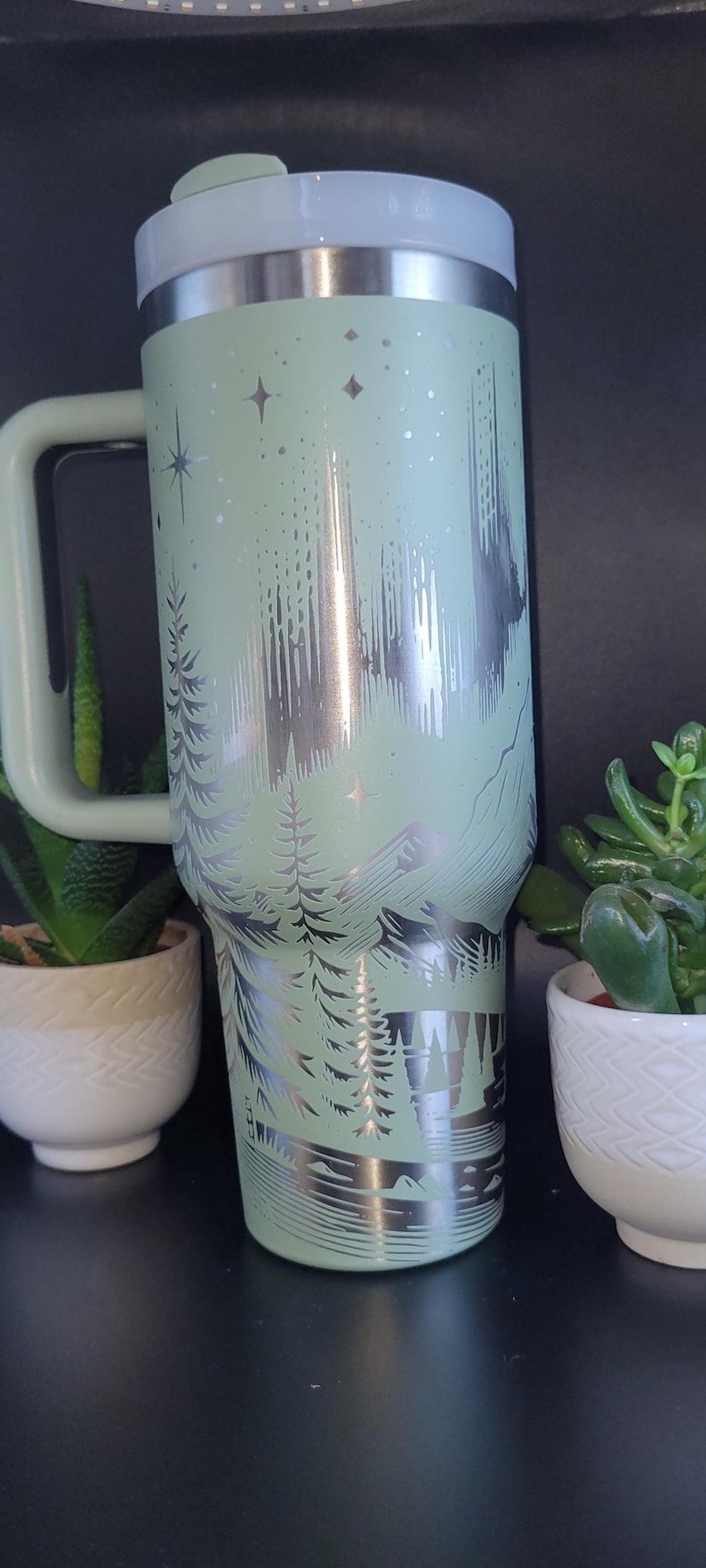 May include: A mint green travel mug with a silver mountain and forest design. The mug has a handle, a white lid, and is surrounded by small potted plants. The mug is approximately 9 inches tall.