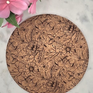 May include: Round cork coaster with a detailed floral design. The coaster features an intricate pattern of flowers, leaves, and butterflies in dark brown against a light brown background. Pink flowers are visible in the upper left corner.