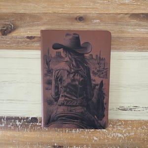 Op de afbeelding: Een bruin notitieboek met een gedetailleerde illustratie van een persoon met een cowboyhoed die door een woestijnlandschap rijdt. Het kunstwerk is in bruin- en zwarttinten, met cactussen en mesas op de achtergrond.