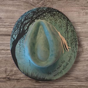 May include: A teal fedora hat featuring a black willow tree design on the brim. The hat has a matching teal crown and a beige tassel. The text "She can make the weeping willows smile" is printed on the brim.