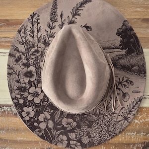 Könnte beinhalten: Ein taupefarbener Cowboyhut mit breiter Krempe und floralem und landschaftlichem Design in Dunkelbraun. Der Hut hat eine Wildleder-ähnliche Textur und einen dekorativen Fransenbesatz.