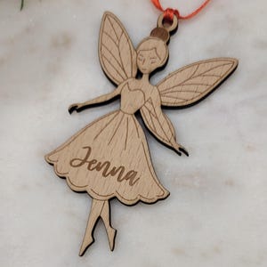 May include: Wooden fairy ornament with the name "Jenna" engraved on the dress. The fairy has detailed wings and is suspended by an orange ribbon. The ornament is made of light-coloured wood with dark edges.