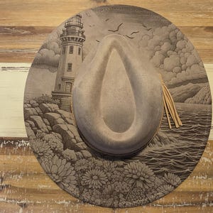 Può includere: Un cappello fedora grigio con un'illustrazione dettagliata di un faro, dell'oceano ed elementi floreali. Il cappello presenta una fascia marrone chiaro e un nappa decorativa. La tesa è ornata da una scena costiera nei toni del grigio e del bianco.