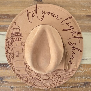 Può includere: Un cappello fedora beige con un design marrone a forma di faro e onde oceaniche. Il cappello presenta la frase "Let your light shine" in corsivo. Il cappello ha una corona color beige e un nappa decorativa.