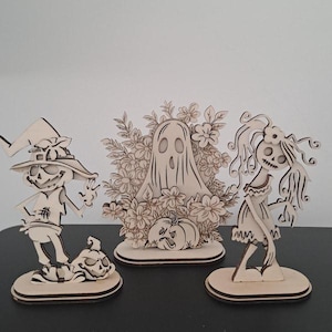 May include: Three wooden Halloween decorations. A scarecrow with a pumpkin, a ghost surrounded by flowers and a skeleton woman. All are laser cut from light-colored wood and stand on oval bases. The decorations are on a black surface.