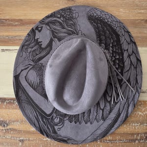 May include: A gray fedora hat with a wide brim featuring a detailed black illustration of an angel with wings. The hat has a tassel detail on the side and is set against a wooden background.