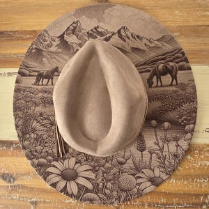 May include: A tan cowboy hat with a detailed landscape design. The brim features a mountain range, horses, and floral elements. The hat has a rounded crown and a decorative tassel.