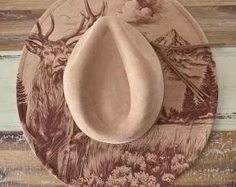 Puede incluir: Un sombrero fedora beige con ala ancha, decorado con una ilustración marrón de un ciervo, montañas y árboles. El sombrero tiene una textura similar al ante y un detalle de cordón de cuero.