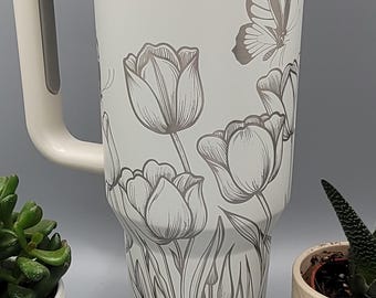 May include: White stainless steel travel mug with a black butterfly and tulip design. The mug has a handle and a lid.