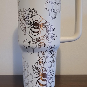 Könnte beinhalten: Ein weißer Edelstahlbecher mit einem schwarzen und goldenen Bienen- und Wabenmuster. Der Becher hat einen Deckel und einen Griff.
