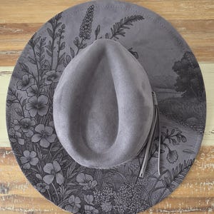 Op de afbeelding: Grijze fedora hoed met een brede rand, met een gedetailleerd bloemen- en landschapsontwerp in zwart. De hoed heeft een bijpassende grijze kwast en staat op een rustiek houten oppervlak. De rand van de hoed heeft een diameter van ongeveer 35.5 cm.