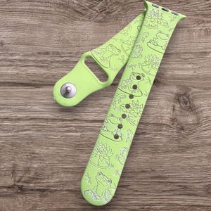 May include: A light green watch strap with a frog and lily pad design. The strap is made of a flexible material and has adjustment holes. The design features detailed illustrations of frogs, lily pads, and dragonflies.