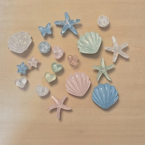 May include: A collection of small decorative resin pieces in pastel colors, including various seashells, starfish, hearts, a butterfly, and round beads. The items feature translucent, glittery, and solid finishes in shades of blue, pink, and green.