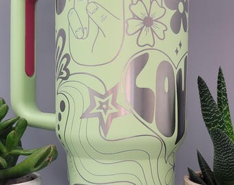 May include: A large, light green travel mug with a white lid and a pink handle accent. The mug is decorated with silver-colored peace signs, flowers, a rainbow, and the word "LOVE".