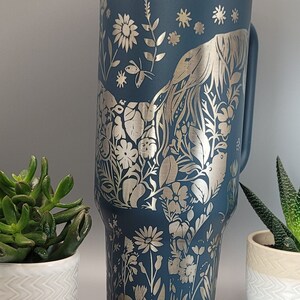 May include: A dark blue stainless steel tumbler with a silver floral design. The design features flowers, leaves, and a bird.