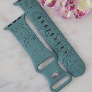 Puede incluir: Dos correas de reloj turquesas con diseños florales y de animales. Una correa presenta un oso y constelaciones, la otra una luna y flores. Las correas están hechas de un material suave y flexible y tienen un cierre plateado.