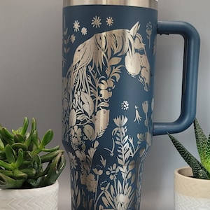 May include: A blue stainless steel travel mug with a silver lid and handle. The mug features a silver metallic design of a horse's head surrounded by flowers.