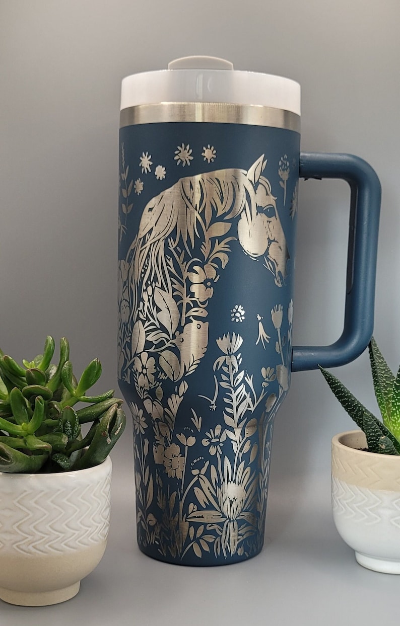 May include: A blue stainless steel travel mug with a silver lid and handle. The mug features a silver metallic design of a horse's head surrounded by flowers.