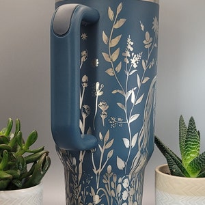 May include: A blue stainless steel travel mug with a silver floral design and a handle. The mug is sitting on a table with a gray background.