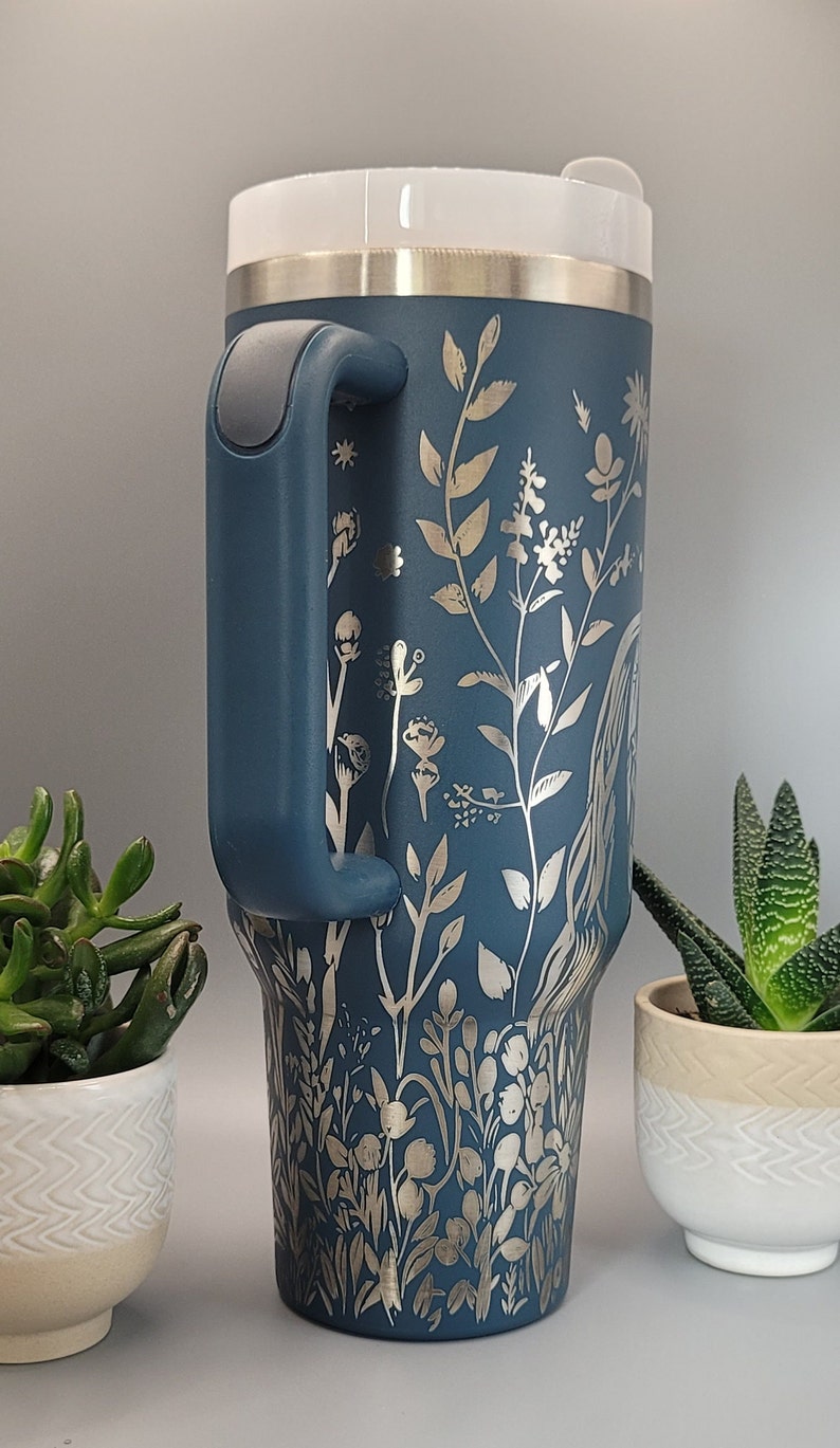 May include: A blue stainless steel travel mug with a silver floral design and a handle. The mug is sitting on a table with a gray background.