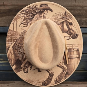 May include: A tan cowboy hat with a detailed illustration of a cowgirl riding a horse. The hat features a light brown suede crown and a wide brim with a sepia-toned graphic of a woman on horseback. The brim has a fringe.