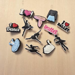 Puede incluir: Una colección de dijes con temática de ballet. Incluye siluetas de bailarinas, un cisne, una zapatilla de ballet y un tutú. Los dijes también muestran las frases "I Dance" e "I Ballet". Los colores son negro, blanco, rosa y azul.