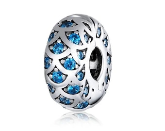 Puede incluir: Charm plateado con un diseño calado y gemas azules incrustadas. El charm ovalado tiene un agujero para ensartar en una pulsera o collar. Las gemas son redondas y están espaciadas uniformemente.