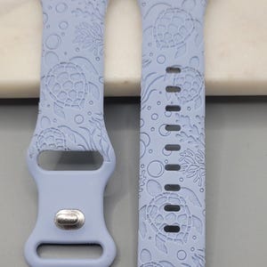 Può includere: Due cinturini per orologio azzurri con un motivo a tartaruga marina. I cinturini sono realizzati in materiale flessibile e presentano un motivo ripetuto di tartarughe, coralli e bolle. Progettato per uno smartwatch.