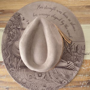 Op de afbeelding: Een beige fedora hoed op een ronde, bedrukte ondergrond. De ondergrond heeft een gedetailleerde illustratie van een vrouw met lang haar en bloemmotieven, samen met de tekst: "Haar kracht werd geboren in stilte, haar moed gevormd door de wind."