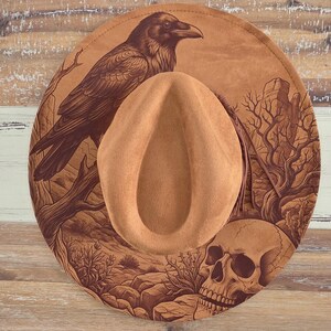 May include: A tan wide-brimmed hat featuring a dark brown laser-engraved design on the brim. The illustration depicts a large raven perched on a branch, a human skull, and desert landscape elements including rocks and sparse vegetation.