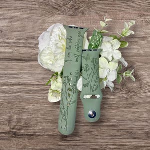 Puede incluir: Dos correas de reloj verde salvia con diseños florales y de mariposas. Una correa tiene el texto "I am he who will sustain you" y "Isaiah 46:4". Las correas se exhiben con flores blancas y vegetación sobre una superficie de madera.