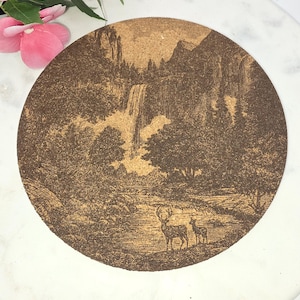 May include: Circular cork coaster featuring a detailed brown illustration of a waterfall, trees, and deer in a natural landscape. The coaster has a neutral colour and a rustic, vintage aesthetic.