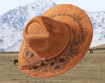 May include: A brown suede cowboy hat with a floral design and the phrase "she is a wildflower" printed on the brim. The hat features a braided band and a tassel. The background shows a mountain range and a grassy field.