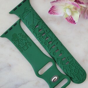 May include: Two green watch straps with engraved floral and mushroom designs. The straps are made of a flexible material and feature a silver clasp. The straps are shown against a background of pink and white flowers.