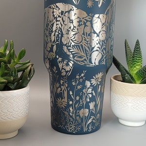 May include: A blue stainless steel tumbler with a silver floral design. The tumbler has a white lid and is perfect for keeping drinks cold or hot.